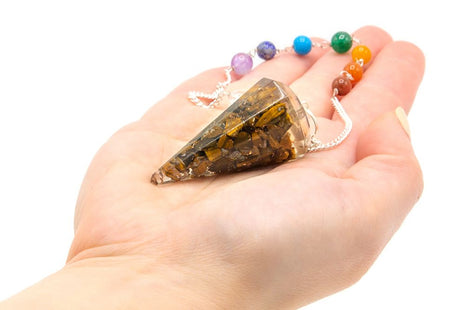 Péndulo de orgonita de ojo de tigre con 7 chakras sostenido en la mano para radiestesia y armonización energética